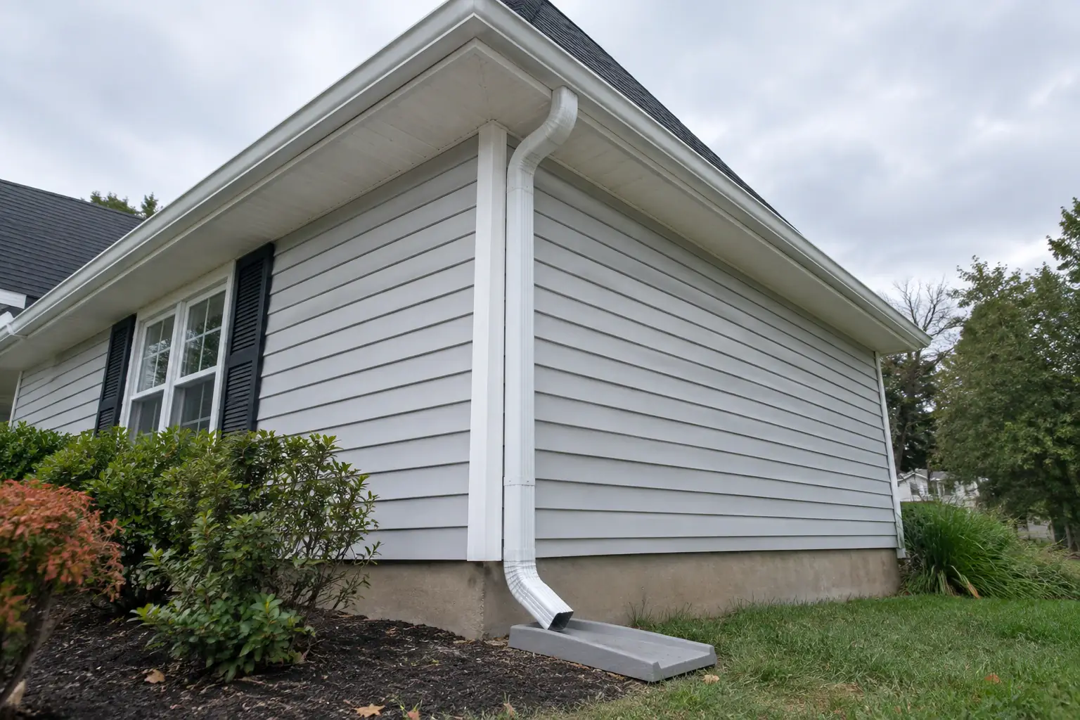 New white gutter along a roof edge with a downspout running down the corner of the house to a splash block.