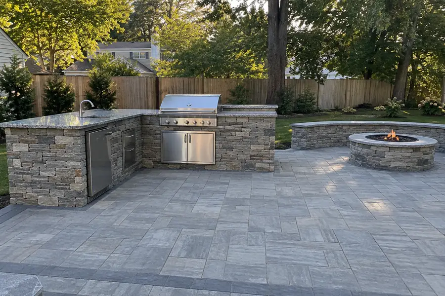 Stone outdoor kitchen island with a grill on a paver patio, a fire pit behind.