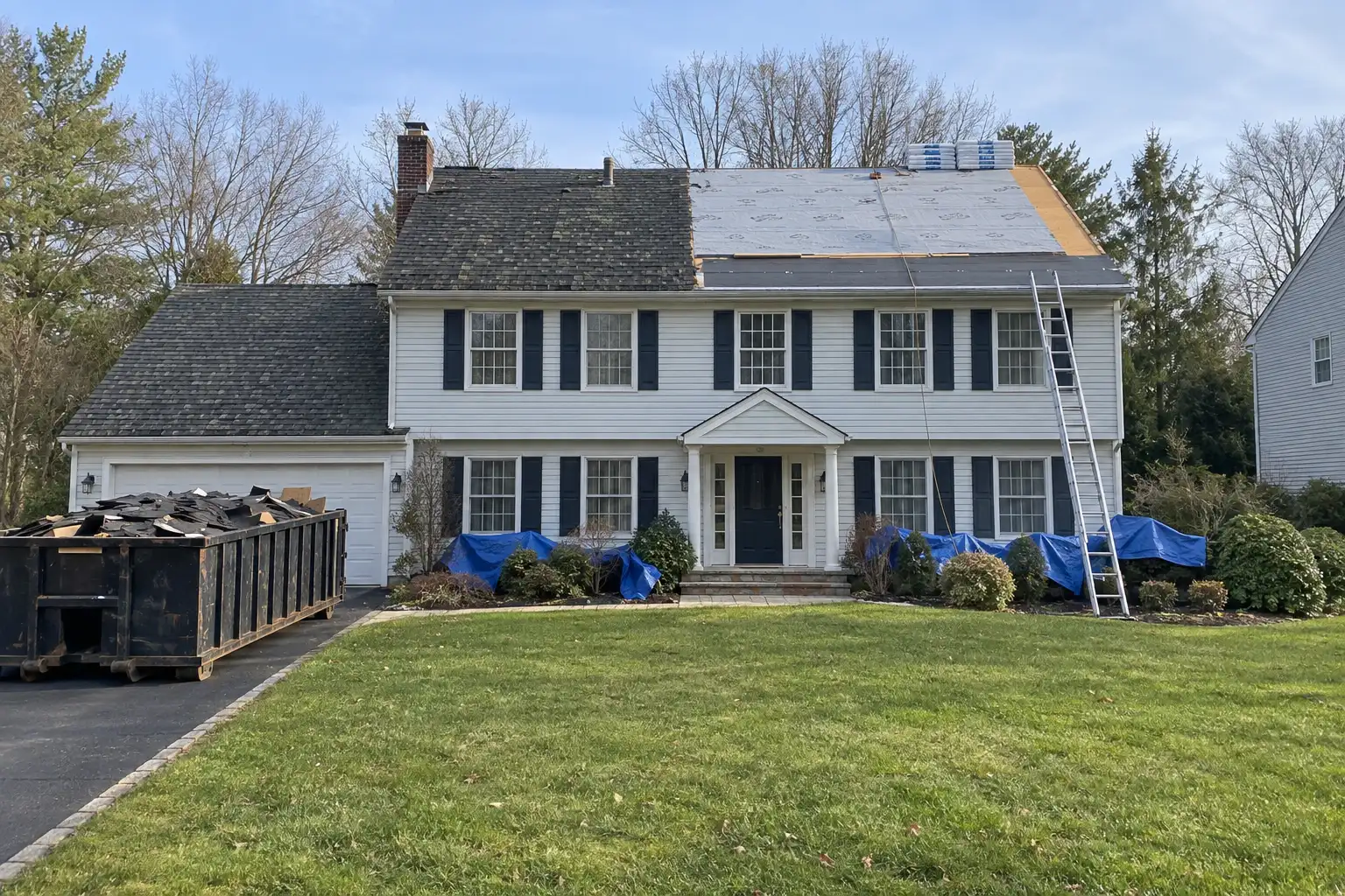 Colonial house mid tear off, half the old shingles removed to the deck and underlayment, a dumpster in the driveway and tarps over the shrubs.