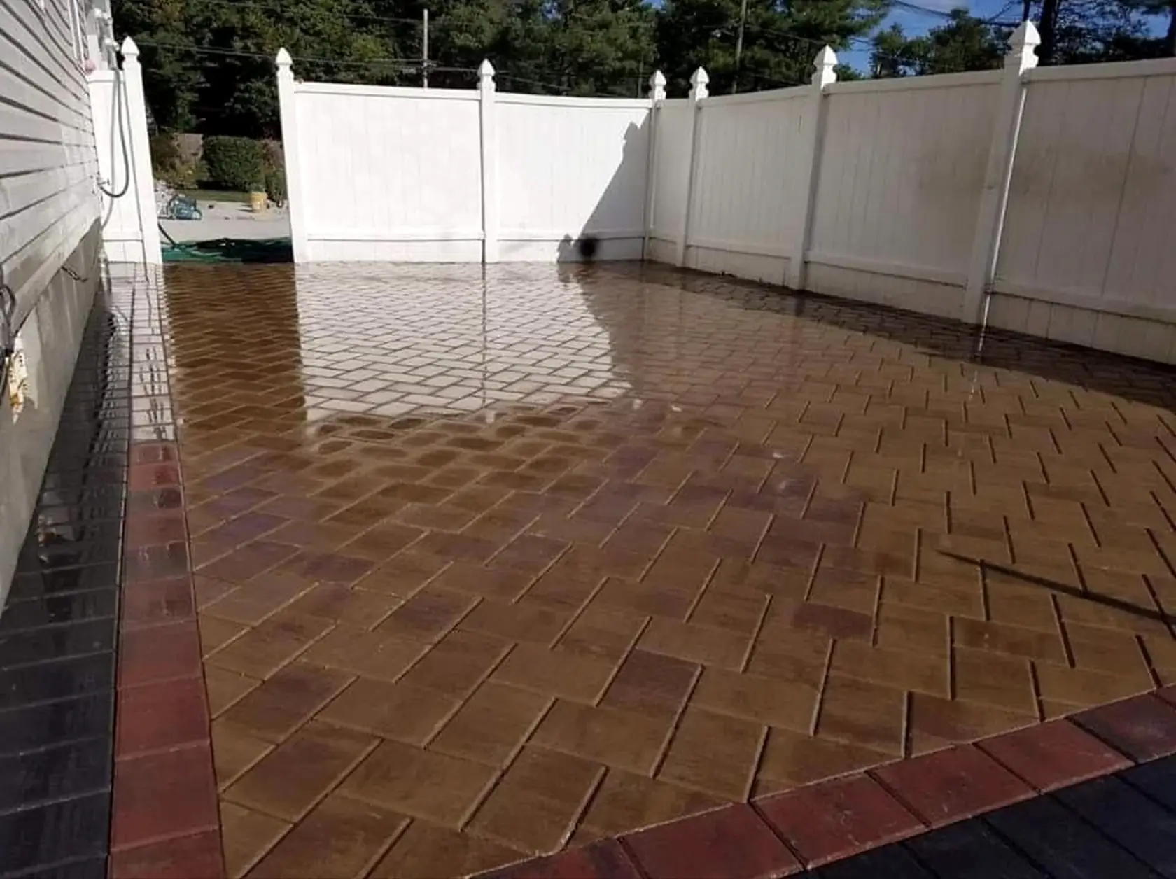 Brick paver patio in a herringbone pattern with a contrasting border and a white fence behind.