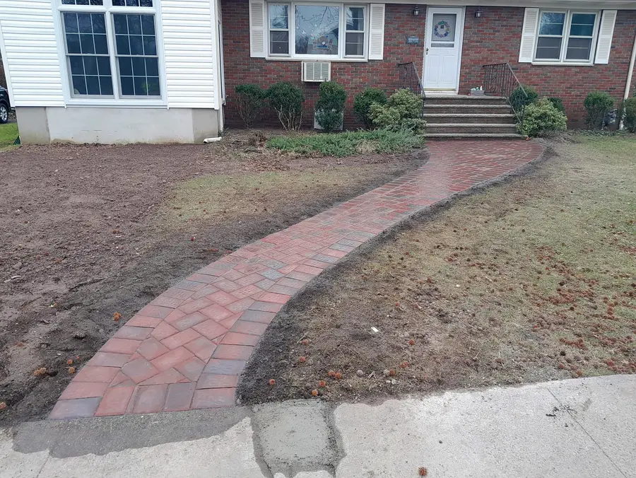 Curved red paver walkway across a lawn toward a brick house.